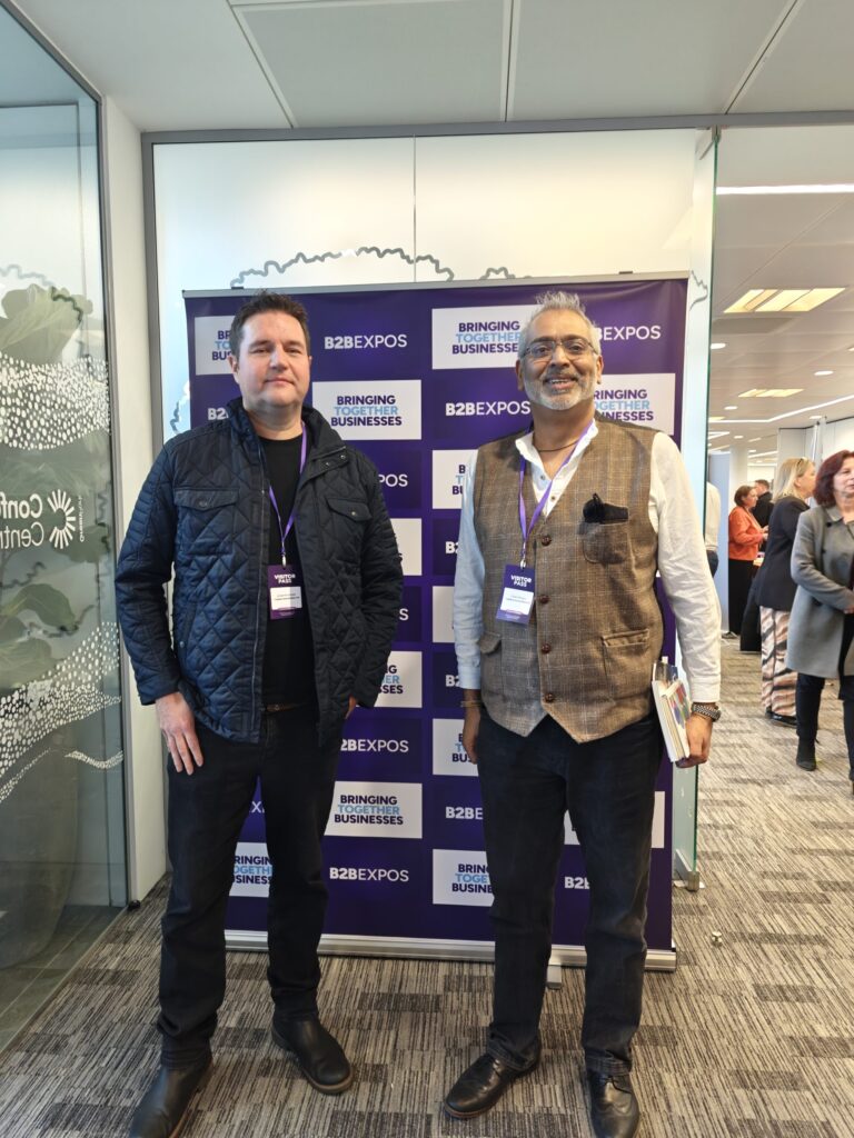 Mark and Vivek -- two business professionals at a B2B Expo event, standing in front of a branded backdrop that reads 'Bringing Together Businesses.' The event is bustling with networking opportunities in a modern venue.